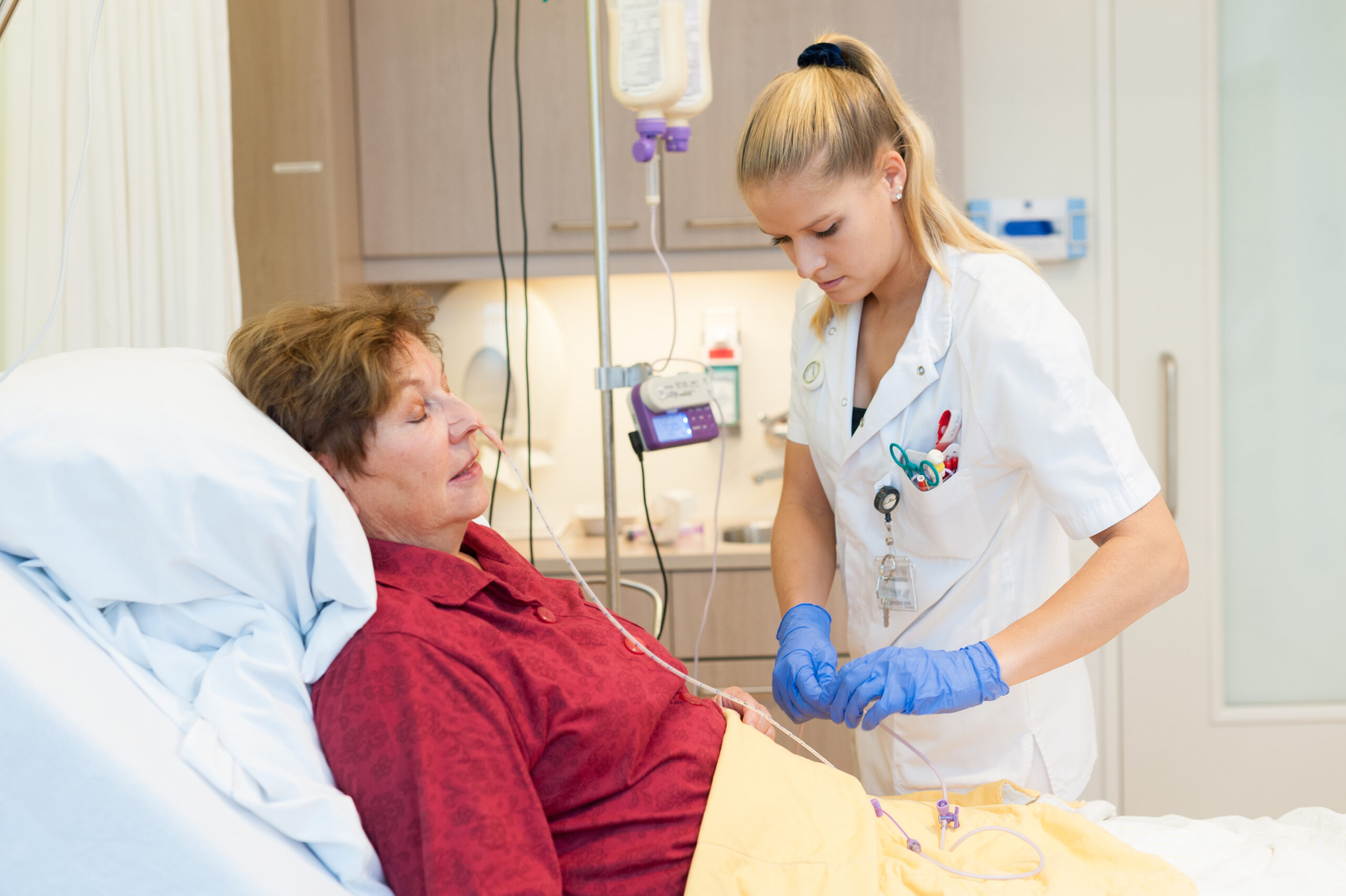 Een patient ligt in een ziekenhuisbed met een sonde in de neus. Een verpleegkundige staat aan het bed en houdt de toediningslijn vast.