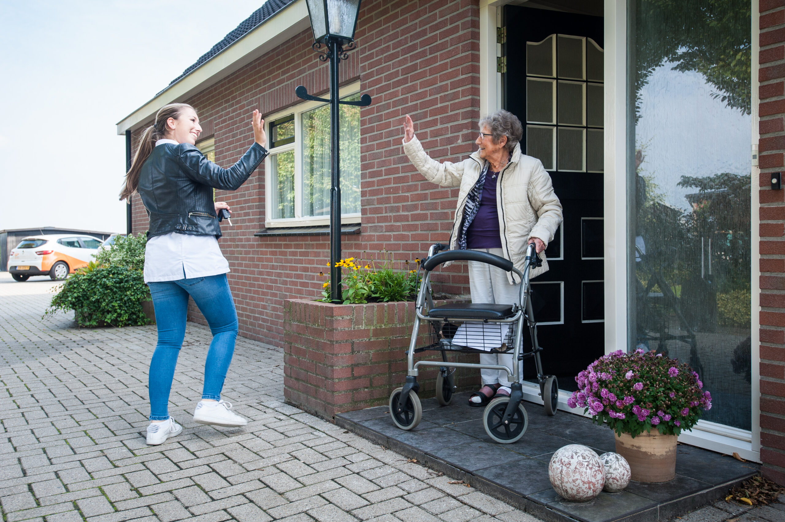 Verpleegkundige en client zwaaien elkaar gedag. De client staat buiten en houdt haar rollator vast.